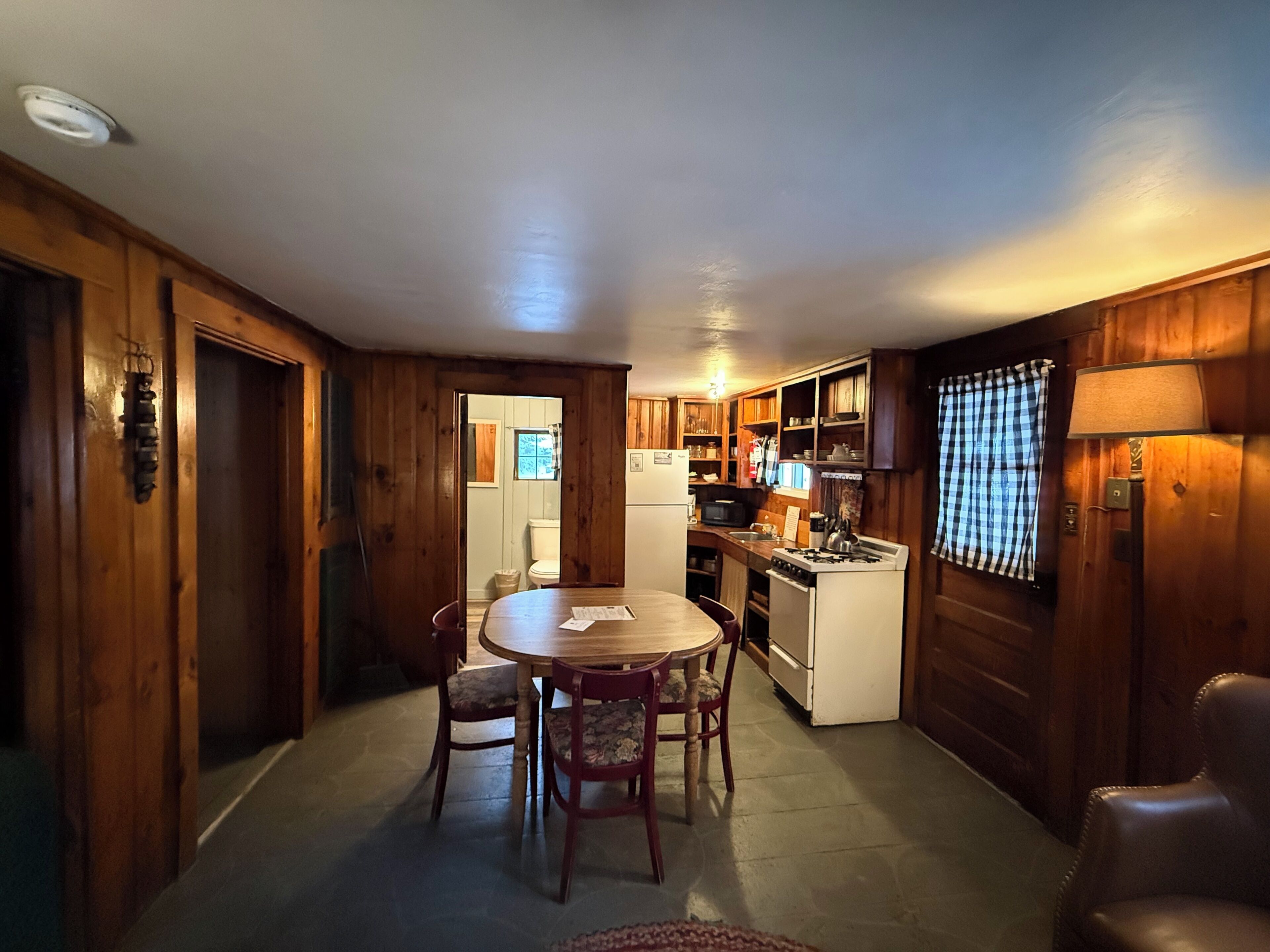 A wooden interior with a dining table, chairs, a kitchen area with a stove and refrigerator, and a window with curtains.