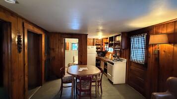 A wooden interior with a dining table, chairs, a kitchen area with a stove and refrigerator, and a window with curtains.