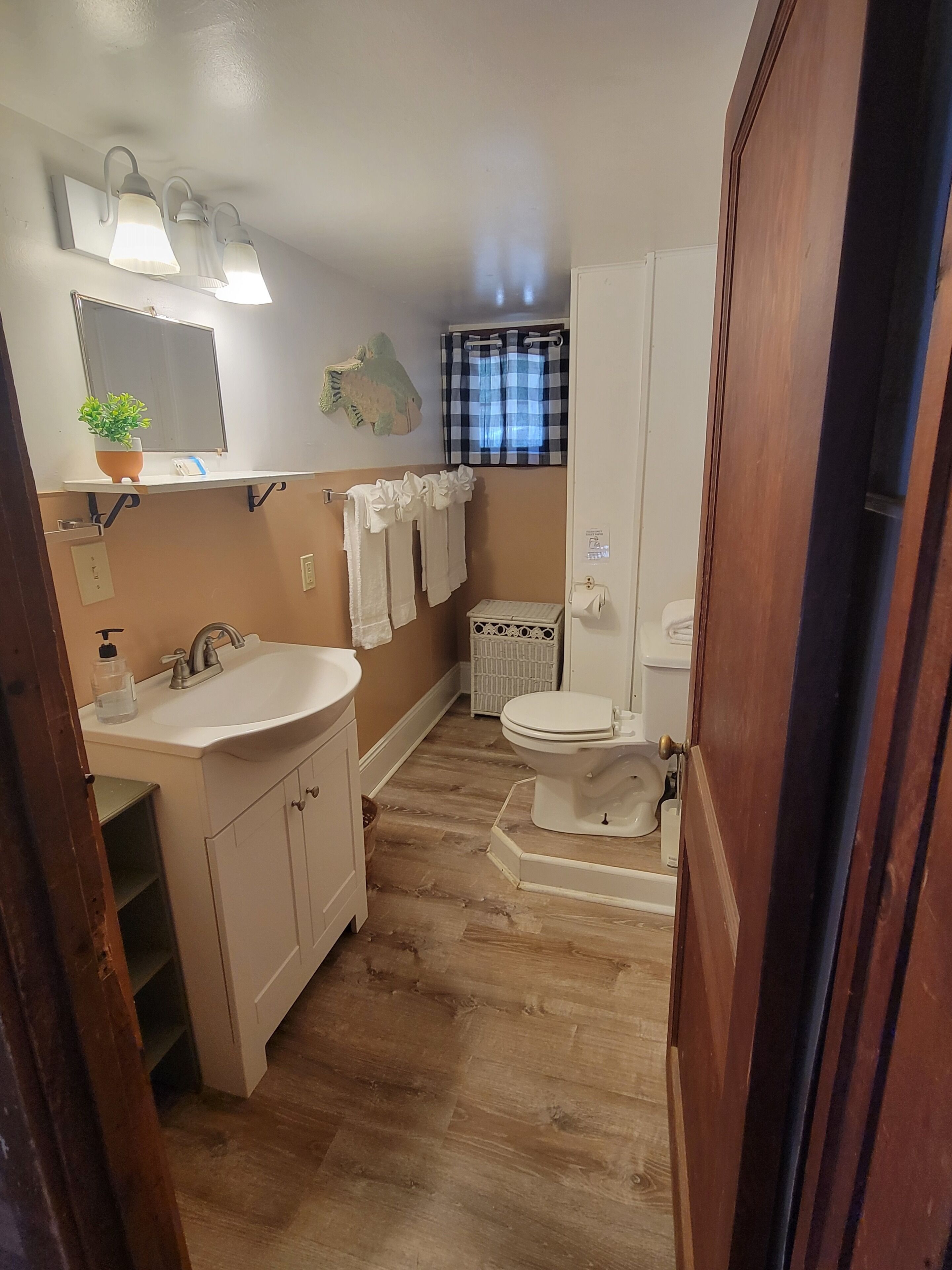 Baño con lavamanos, inodoro y zona de ducha. Hay un espejo sobre el lavamanos y un perchero con toallas.