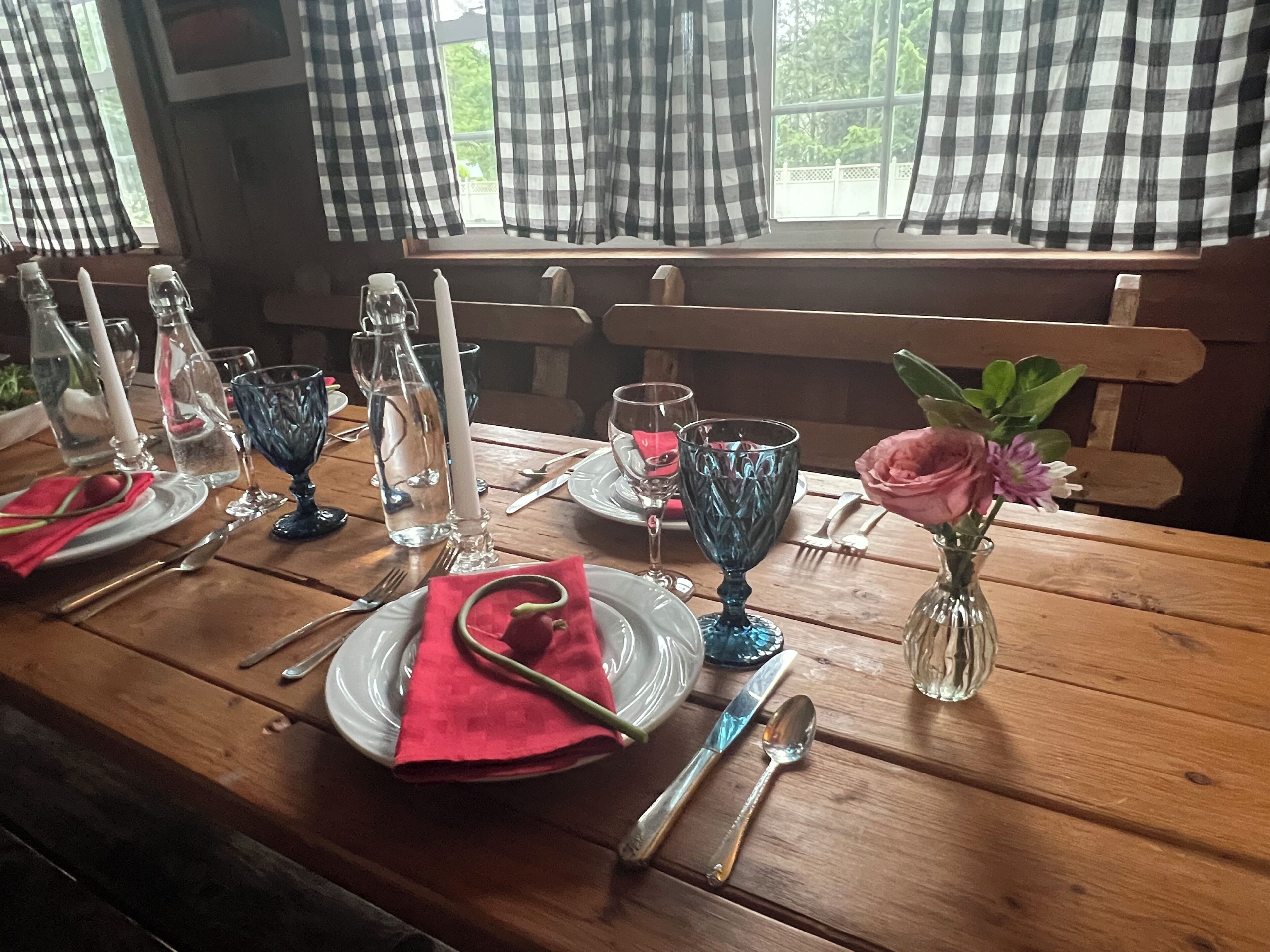 Una mesa de comedor de madera dispuesta con platos, cubiertos, vasos y un florero con flores.