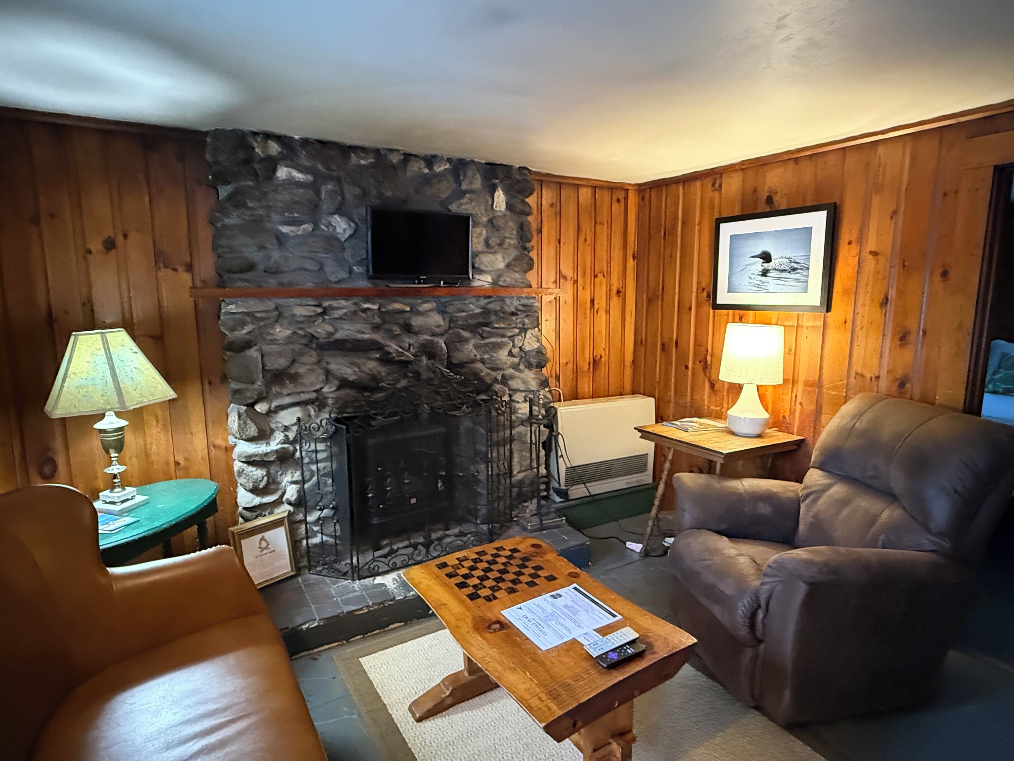 A cozy living room with a stone fireplace, wooden walls, a television, a framed picture, a chessboard, and leather furniture.