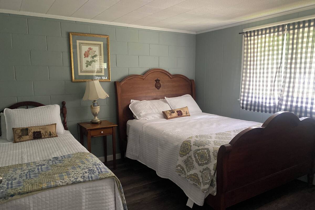 A bedroom with two beds, a nightstand, a lamp, a framed picture, and a window with a checkered curtain.
