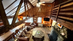 A rustic cabin interior with a wooden ceiling, a stone fireplace, a wooden dining table, leather sofas, and a television mounted on the wall. - Cottage Place on Squam Lake (Holderness)