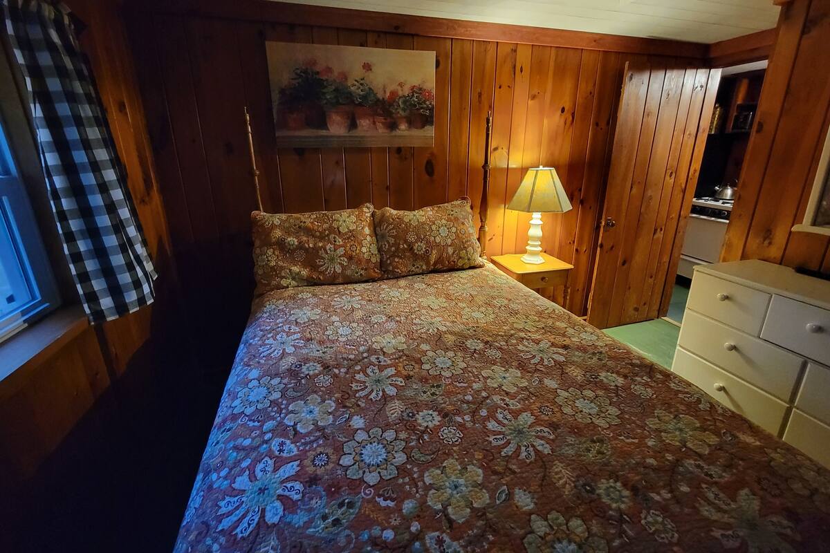A bedroom with a wooden paneled wall, a bed with floral bedding, a nightstand with a lamp, and a dresser.