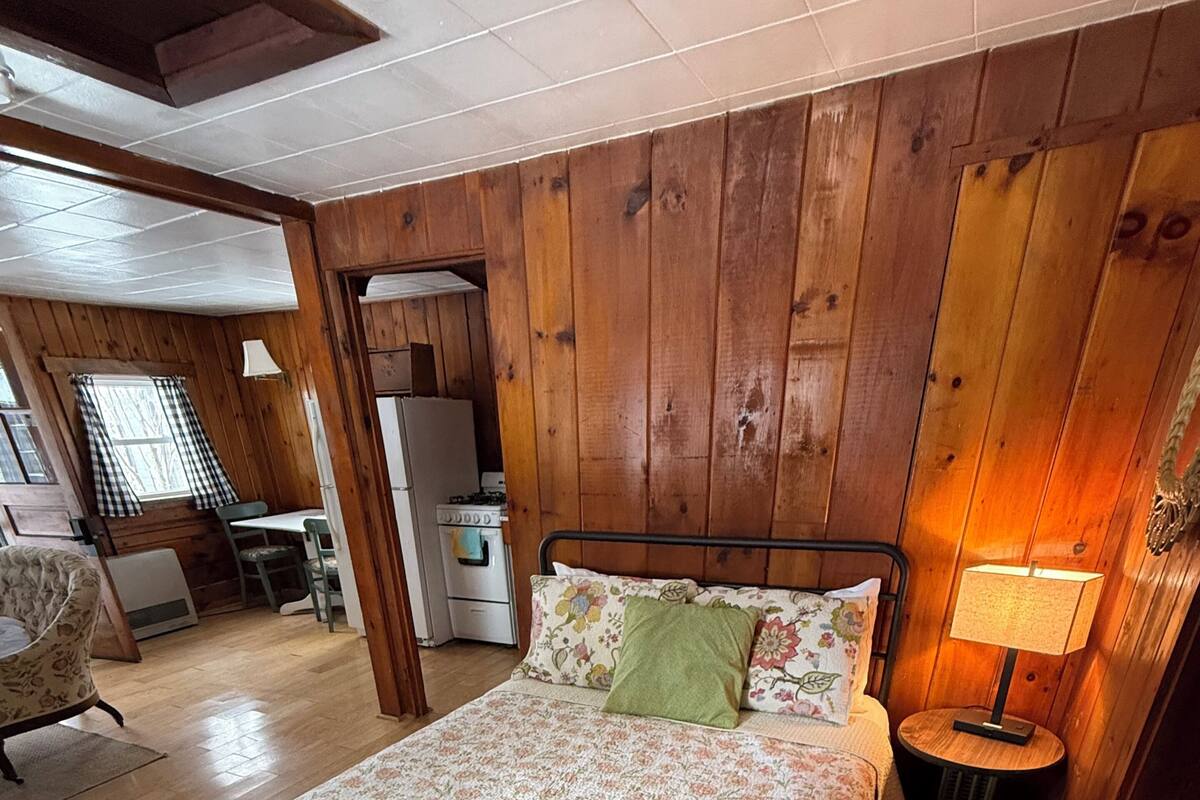 A wooden-paneled bedroom with a bed, a nightstand, a lamp, a chair, a table, and a kitchenette.
