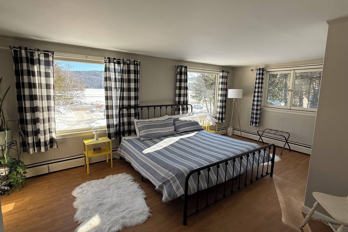 A bedroom with a metal bed frame, a fluffy rug, a bedside table with a lamp, and a view of a snowy landscape through large windows.