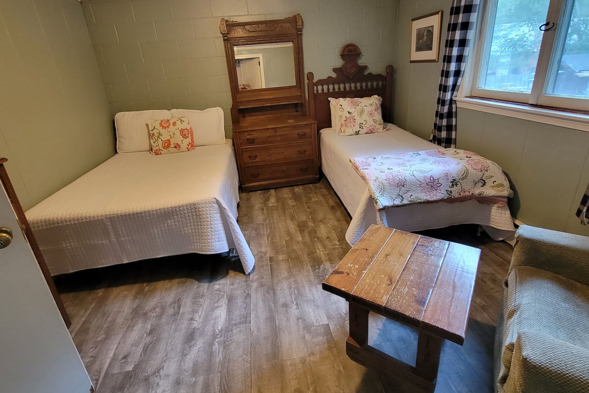 A room with two beds, a wooden dresser, a small table, and a window with curtains.
