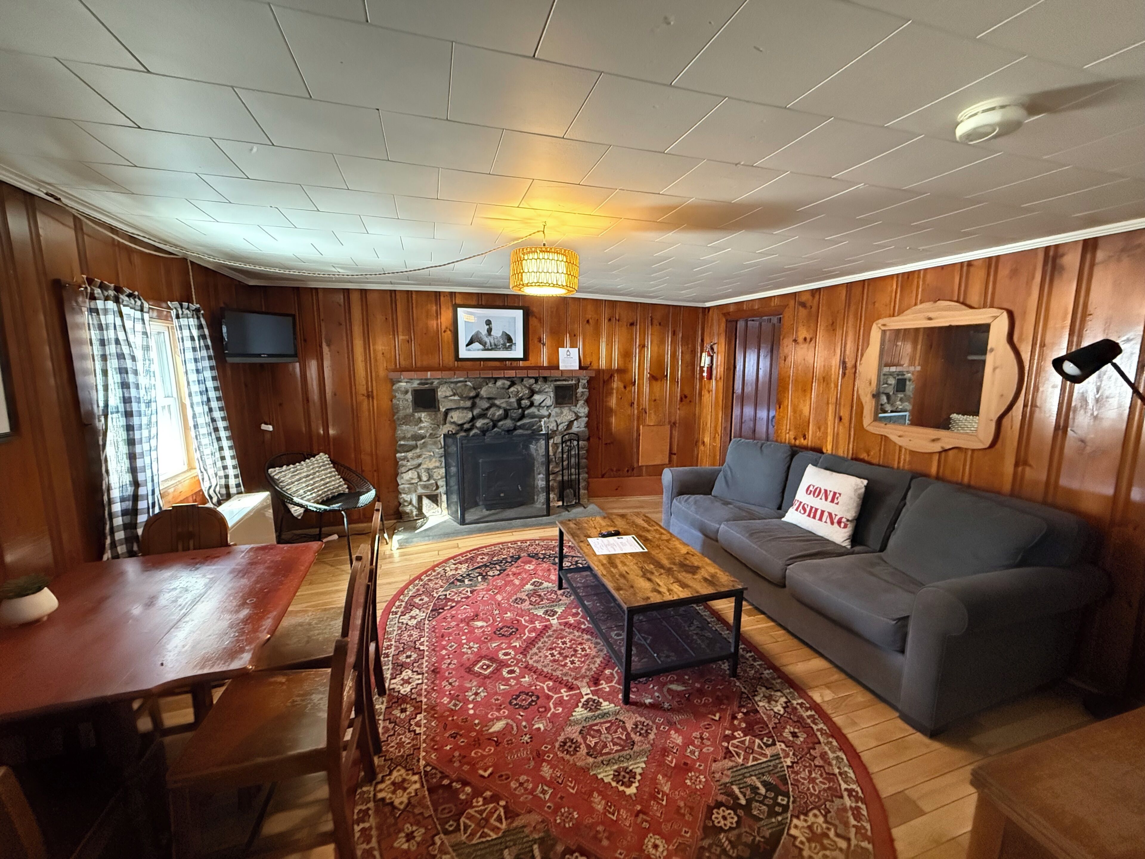 A cozy living room with a stone fireplace, wooden walls, a grey sofa, a red patterned rug, and a dining area with a wooden table and chairs.