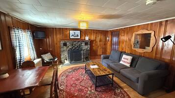A cozy living room with a stone fireplace, wooden walls, a grey sofa, a red patterned rug, and a dining area with a wooden table and chairs.