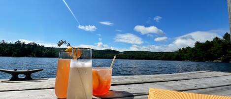 A wooden deck with two glasses of beverages, a small table, and a view of a lake and forest.