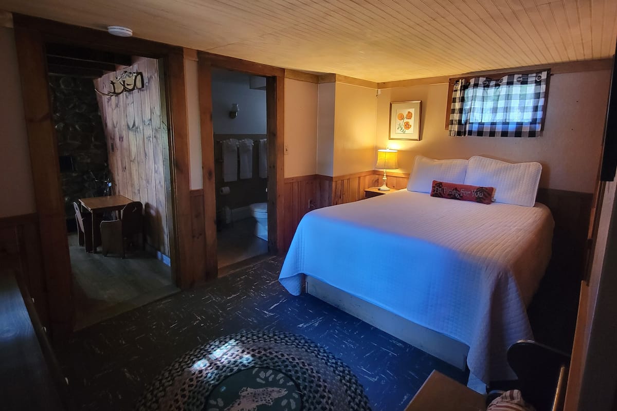 A hotel room with a bed, a small table, and a chair. There is a bathroom visible through an open door.
