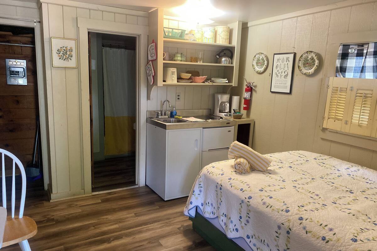 A compact room with a bed, a small kitchenette, a wooden floor, and a door leading to another room.