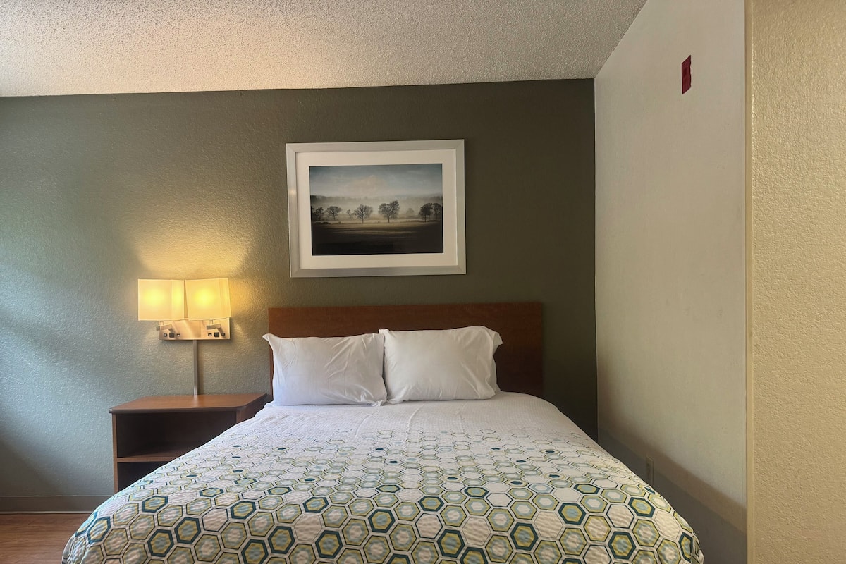 A neatly made bed with a patterned bedspread, a framed picture on the wall, and a bedside lamp.