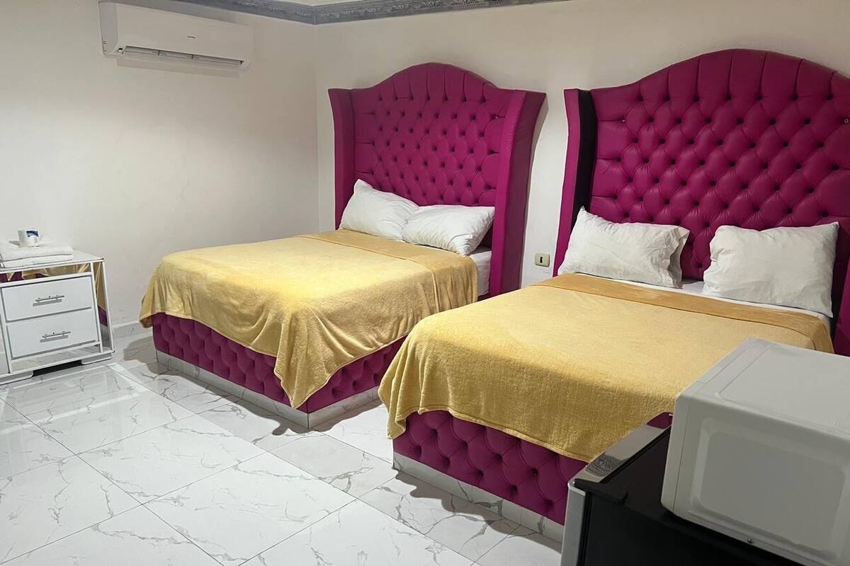 Habitación de hotel con dos camas, cada una con cabecero rosa y ropa de cama amarilla. Hay una mesita de noche blanca con cajones, un equipo de aire acondicionado blanco en la pared y un microondas blanco sobre una encimera negra.