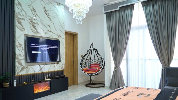 A modern hotel room with a flat-screen TV mounted on a marble wall, a hanging chair with cushions, a bed with a Chanel blanket, and a large window with curtains. - ABOT Luxury Homes (Lekki, Lagos State)