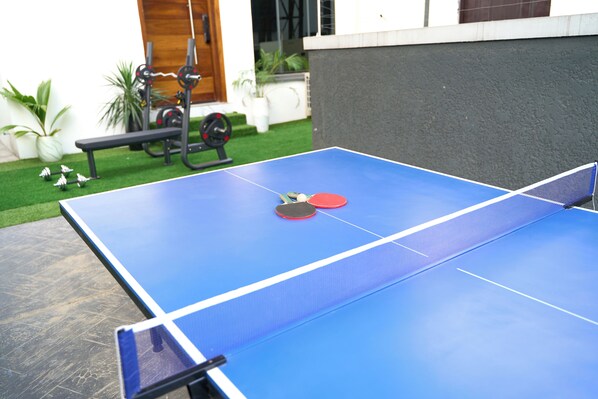 A blue ping pong table with two paddles and a ball in the center, set against a backdrop of a house and outdoor gym equipment. - ABOT Luxury Homes (Lekki, Lagos State)