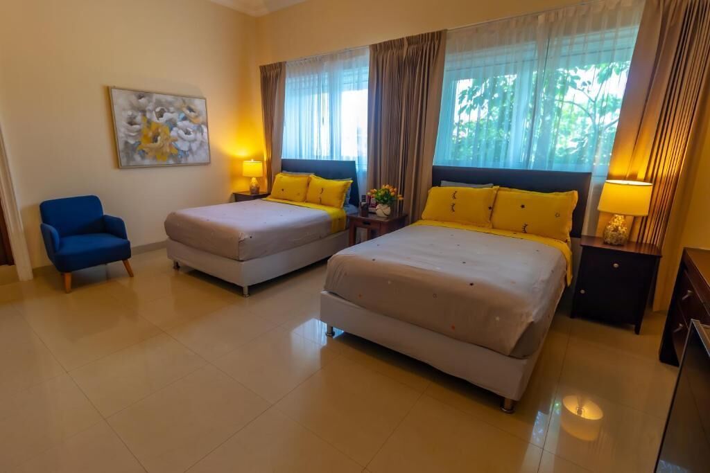 A hotel room with two beds, a blue armchair, and a painting on the wall.