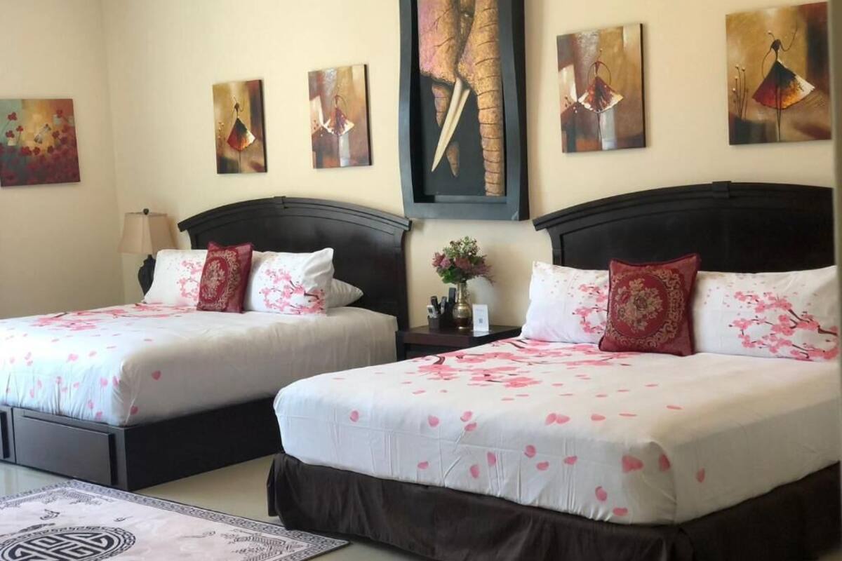 Une chambre d’hôtel avec deux lits, chacun recouvert d’une courtepointe blanche et agrémenté de coussins à motifs floraux roses. Des tableaux sont accrochés au mur au-dessus des lits, et une petite table avec un vase de fleurs se trouve entre les deux.