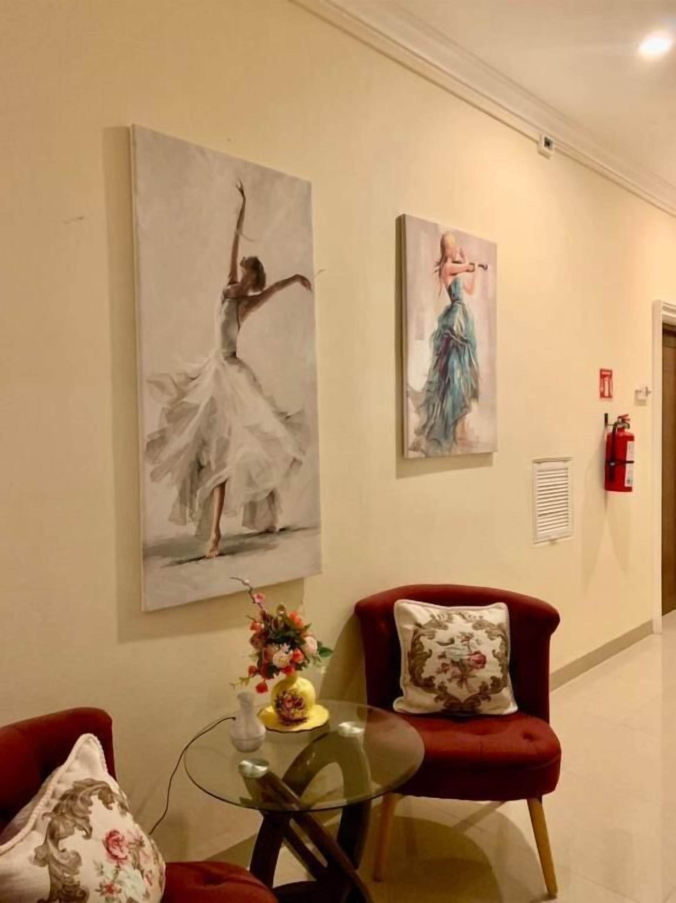 A hallway with two red armchairs, a glass table with a vase and flowers, and two large wall paintings of dancers.