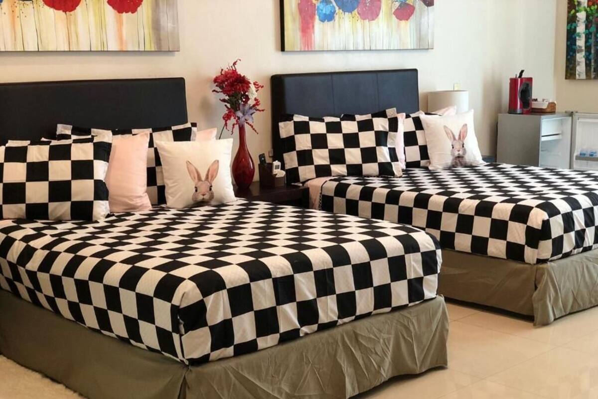 Une chambre d’hôtel avec deux lits, chacun recouvert d’une couvre-lit à carreaux noirs et blancs, assortie aux coussins. On y trouve une table de chevet avec un vase de fleurs et une lampe.