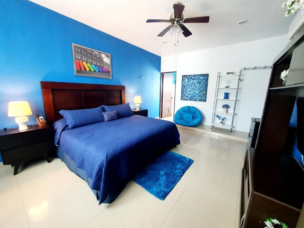 Quarto com colcha azul, ventilador de teto, mesa de cabeceira com abajur, poltrona de feijão mágico azul e televisão em suporte de madeira.