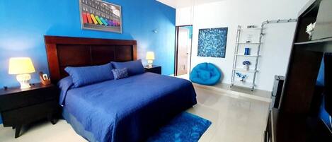 Quarto com colcha azul, ventilador de teto, mesa de cabeceira com abajur, poltrona de feijão mágico azul e televisão em suporte de madeira.