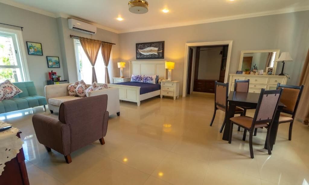 A spacious hotel room with a bed, a sofa, a dining table with chairs, and a small kitchen area.