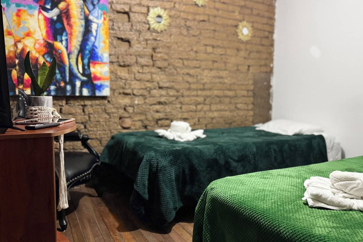 Ein Zimmer mit Backsteinwand, zwei Betten mit grüner Bettwäsche, einem Schreibtisch mit einem Laptop und einem farbenfrohen Gemälde.