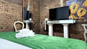 A room with a brick wall, a bed with a green bedspread, a white chair, a white vanity with a mirror, a flat-screen TV, and a painting on the wall. - Casa Cordoba (Bogotá)