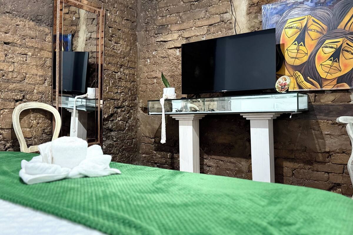 Ein Zimmer mit Backsteinwand, einem Bett mit grüner Bettdecke, einem weißen Stuhl, einer weißen Kommode mit Spiegel, einem Flachbildfernseher und einem Bild an der Wand.