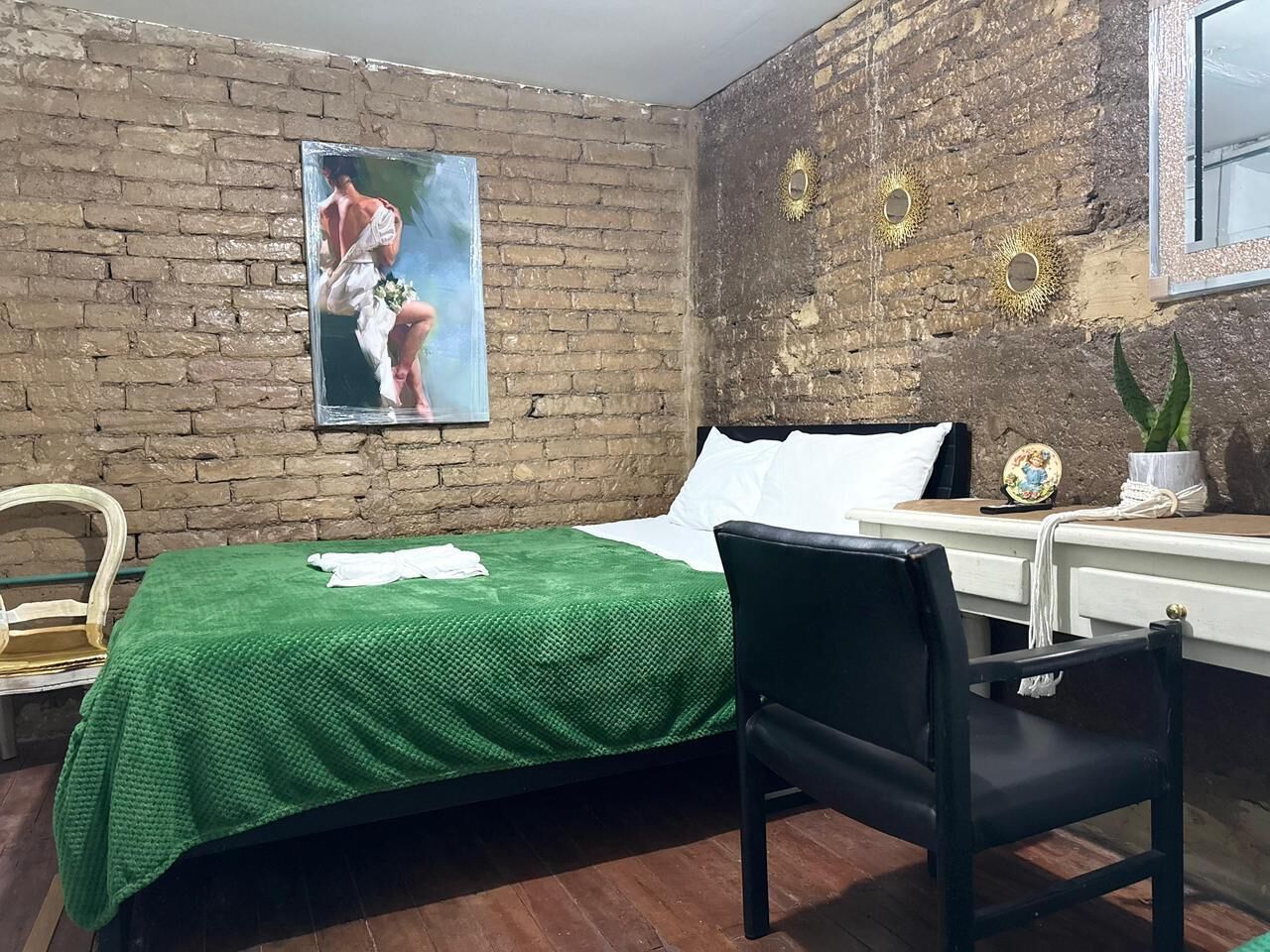 A room with a brick wall, a bed with a green bedspread, a black chair, a desk with a globe, and a picture on the wall.