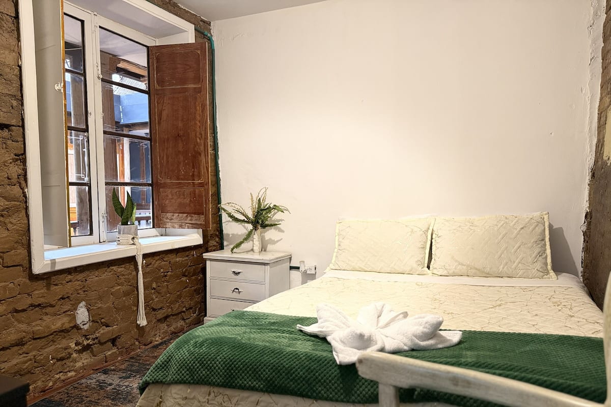 Ein Schlafzimmer mit Bett, Fenster, einem kleinen Nachttisch mit Pflanze und einer Ziegelwand.