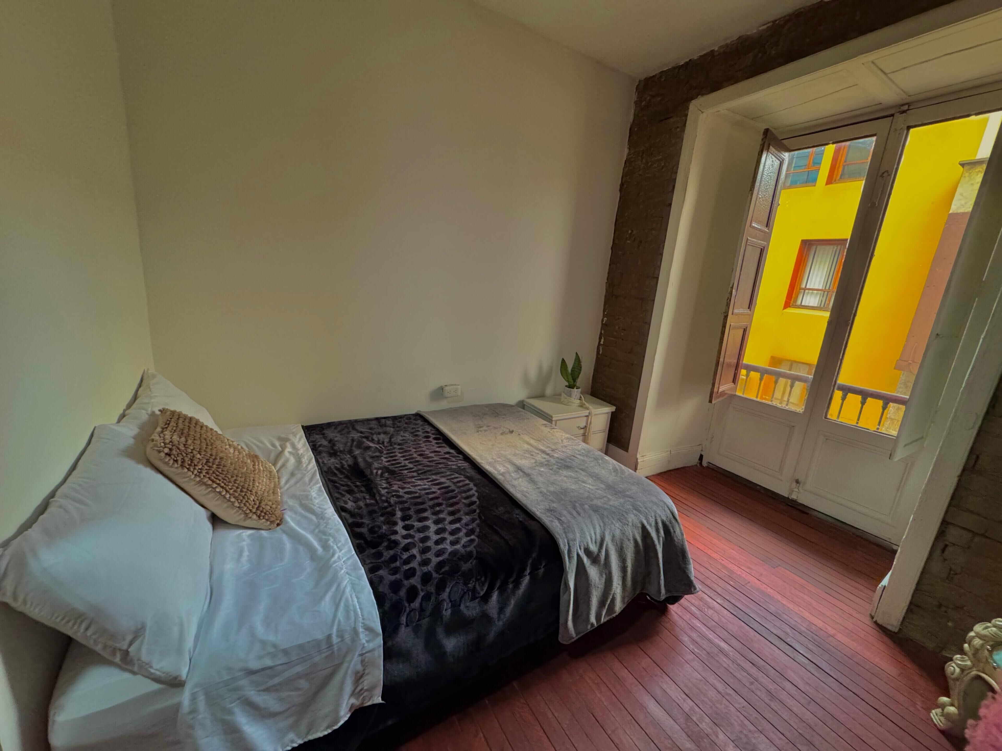 A bedroom with a bed, a bedside table, a potted plant, and a view of a yellow building through a window.