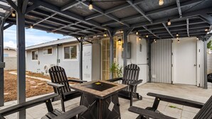 En udendørs terrasse med bord og stole, et overdækket område med synlige bjælker, og udsigt til en bygning i baggrunden.