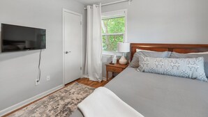 A bedroom with a bed, a TV mounted on the wall, a window with curtains, a bedside table with a lamp, and a rug on the floor.