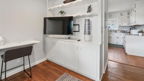 A compact kitchen with white cabinetry, a built-in TV, and a small dining area with a chair.
