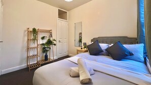 A hotel room with a large bed, a bedside table with a lamp, a wooden plant stand, and a door leading to another room. - Spacious, Cosy & Modern Home w/ Parking , Wi-Fi and Garden (coventry west midlands)