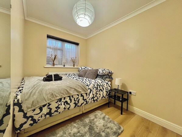 Un dormitorio con cama, una mesita, una lámpara, una ventana con cortinas e iluminación en el techo.