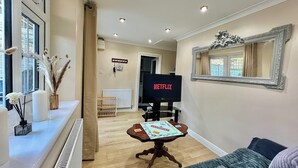 Una sala de estar con un televisor mostrando 'NETFLIX', una mesita con un juego de mesa y un espejo en la pared.