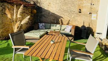 An outdoor seating area with a wooden table and chairs, a cushioned sofa, and a stone wall backdrop.