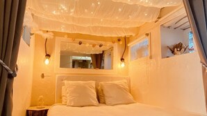 Una cama con sábanas blancas, una mesita de noche, un espejo y luces colgantes en el techo.