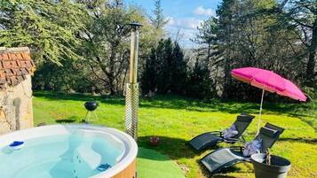 A hot tub in a garden with a brick wall, a pergola, and lounge chairs under a pink umbrella.