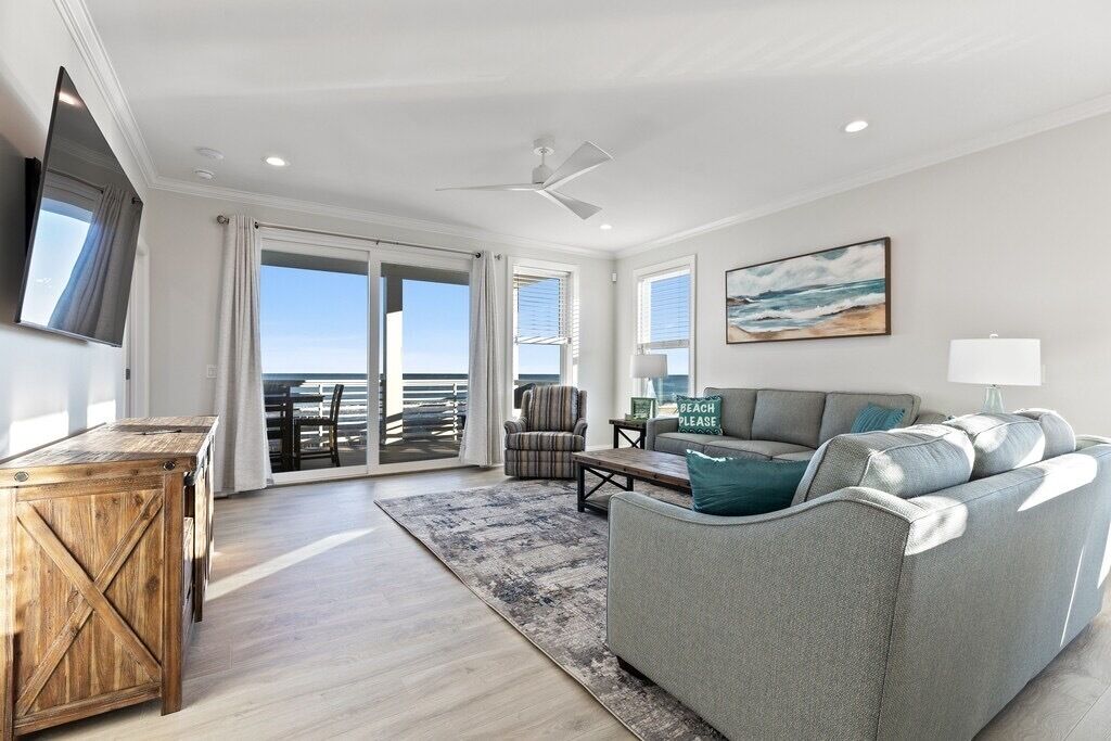 A spacious living room with a large grey sofa, a wooden coffee table, and a TV mounted on the wall. There is a sliding glass door leading to a balcony with a view of the ocean.