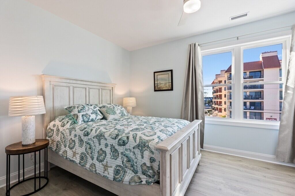A bedroom with a bed, two bedside lamps, a nightstand, a window with curtains, and a framed picture on the wall.