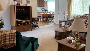 Ett vardagsrum med en tv, en grön soffa, ett träskåp, en matplats med ett bord och stolar samt en mönstrad soffa.