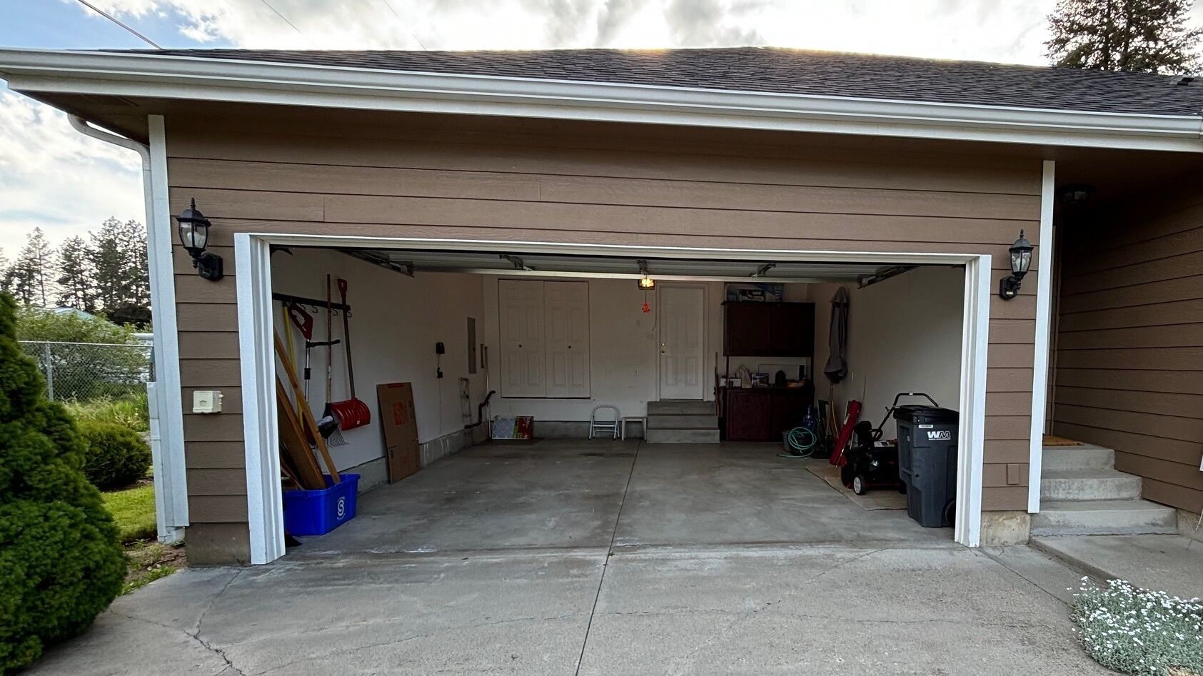 A garage with a concrete floor, a white door, and a small step leading up to the garage door. There are two light fixtures on the wall, a blue trash bin, and a red mailbox.