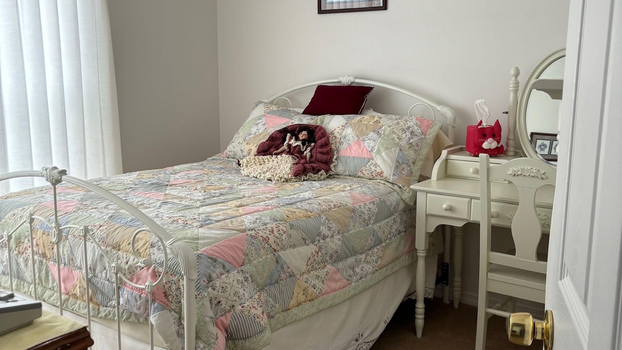 A neatly made bed with a floral quilt, a bedside table with a mirror, and a dresser with a small red toy.
