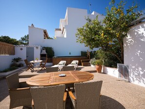 Une terrasse avec une table ronde en bois et des chaises en osier, un espace barbecue et un bâtiment blanc doté d’un toit en pente.