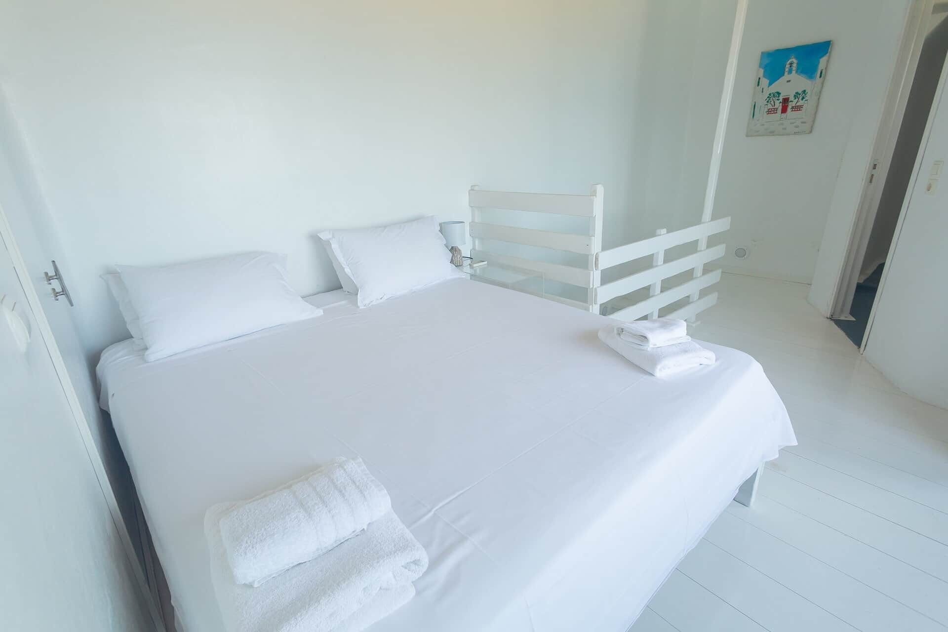 A white bed with pillows and a folded towel, a white headboard, and a painting on the wall.