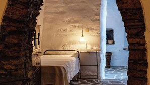 Una habitación rústica y estrecha con una cama, una pequeña mesa con una lámpara y suelo de piedra.
